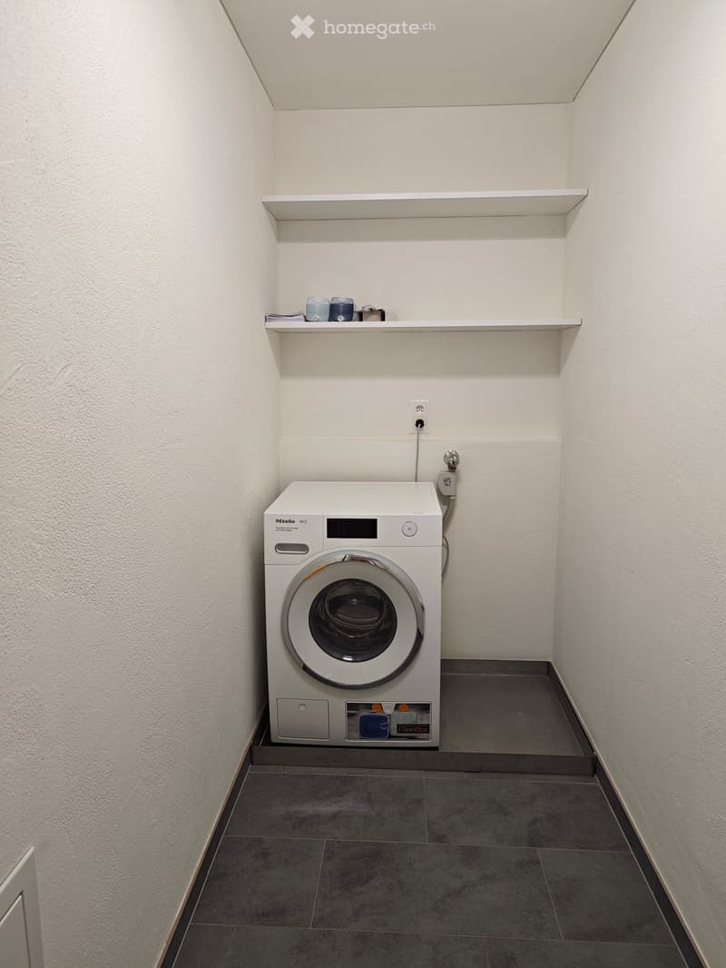 A small, clean utility room with a white washing machine, a built-in shelf holding cleaning supplies, and a tiled floor.