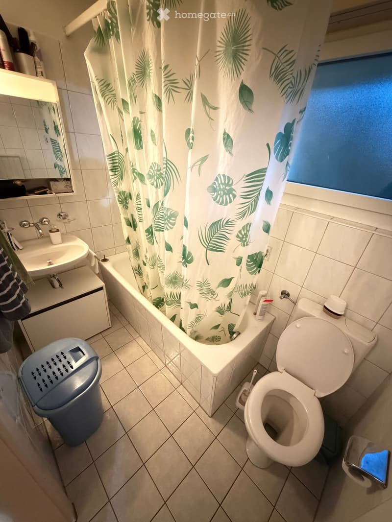 Bathroom with a bathtub, toilet, sink, and mirror. Features a green leaf-patterned shower curtain and tiled walls and floor.