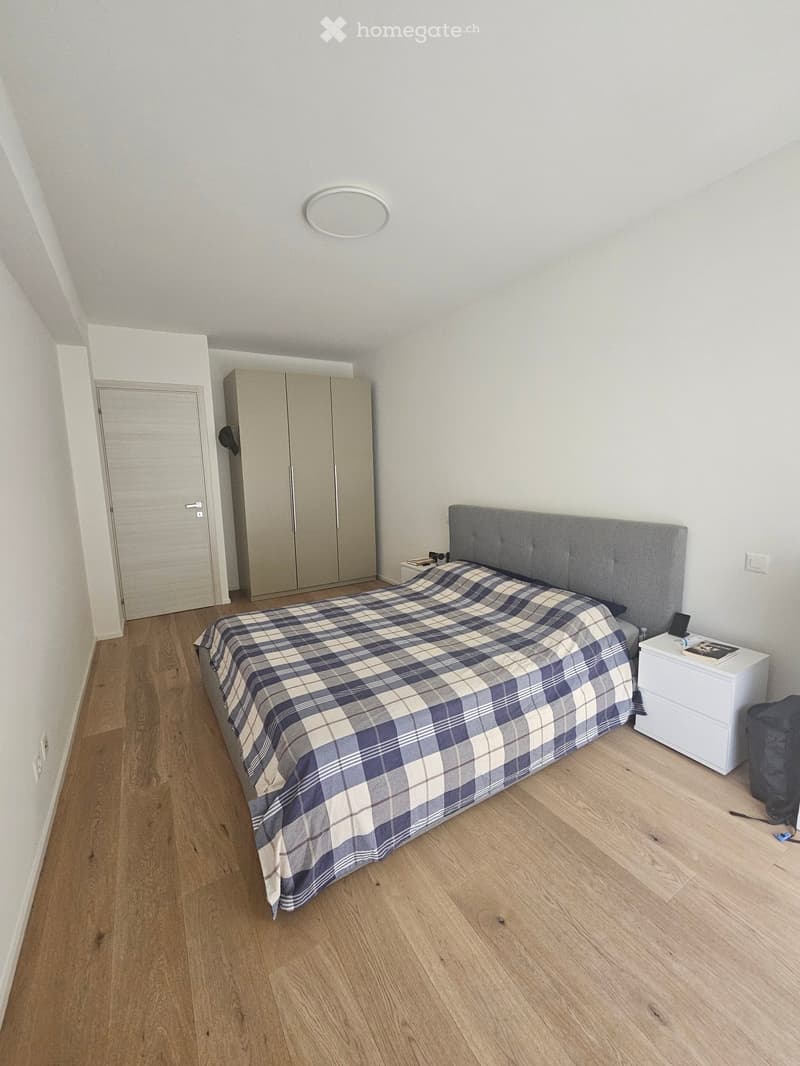Modern bedroom with wooden flooring, a large bed with a grey headboard and plaid duvet, a white nightstand, and a wardrobe with mirrored doors.