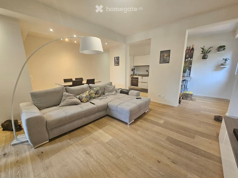 Modern living room with a grey sectional sofa, floor lamp, dining area with chairs, and a small kitchen area with cabinets and an oven. Shelves hold bottles and plants, and a framed picture adorns the wall.
