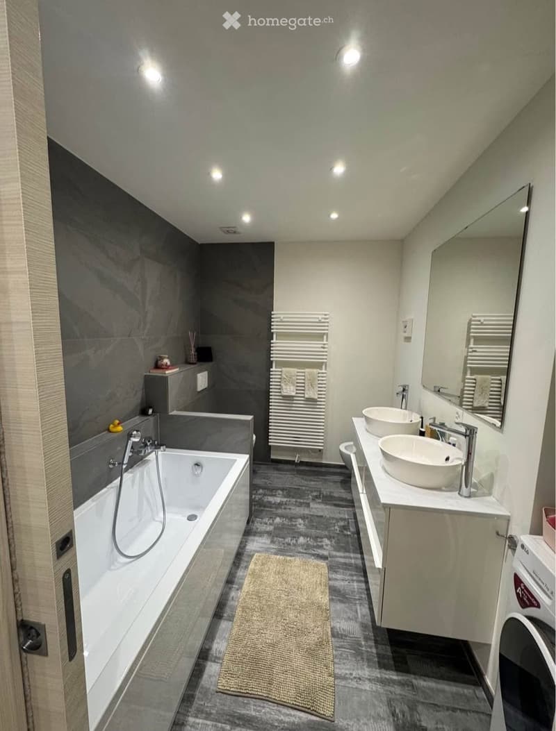 Modern bathroom with a bathtub, double sink, large mirror, radiator, and a small washing machine. The walls are tiled in dark grey, and the floor is dark grey with a beige rug.