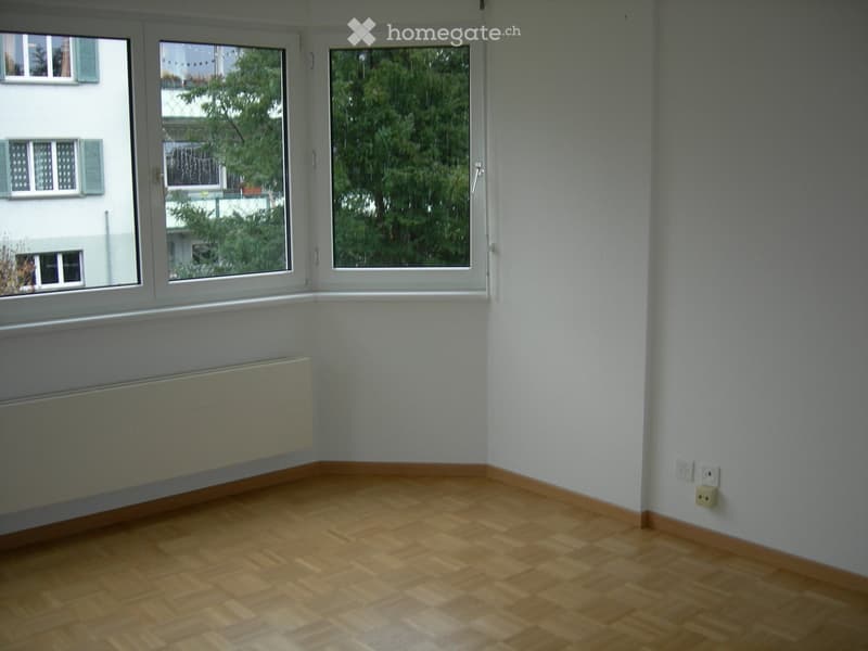 Empty room with wooden flooring, white walls, large windows, radiator, and electrical outlets.