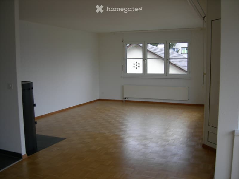 Empty room with wooden flooring, white walls, and large windows letting in natural light. There is a radiator below the windows and a door visible on the left side.