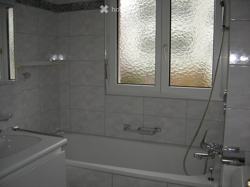 Modern bathroom with a bathtub, shower hose, and a cabinet. The walls are tiled, and there is a window with frosted glass above the bathtub.