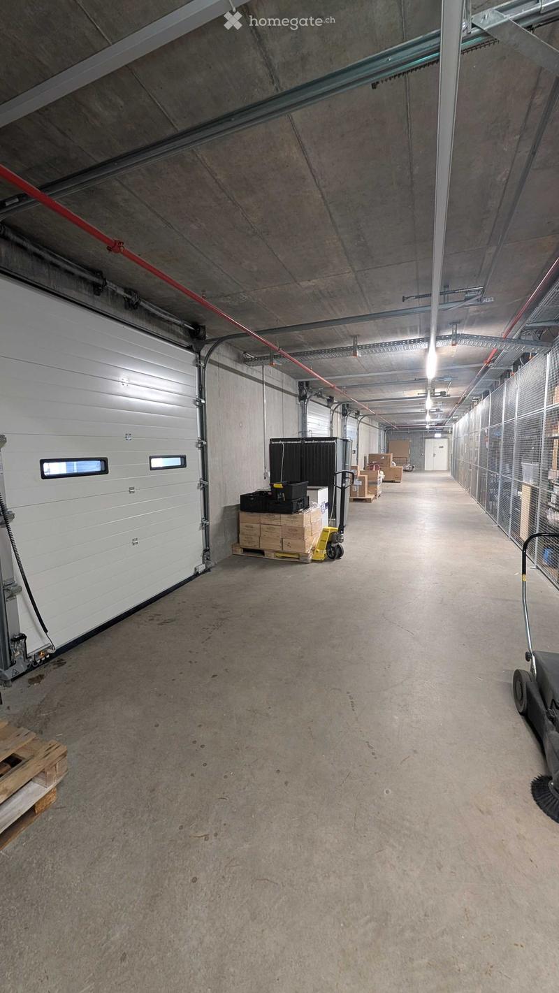 A long, wide concrete corridor with a high ceiling, industrial lighting, and a large garage door on the left. Storage items and a pallet are visible near the garage door.