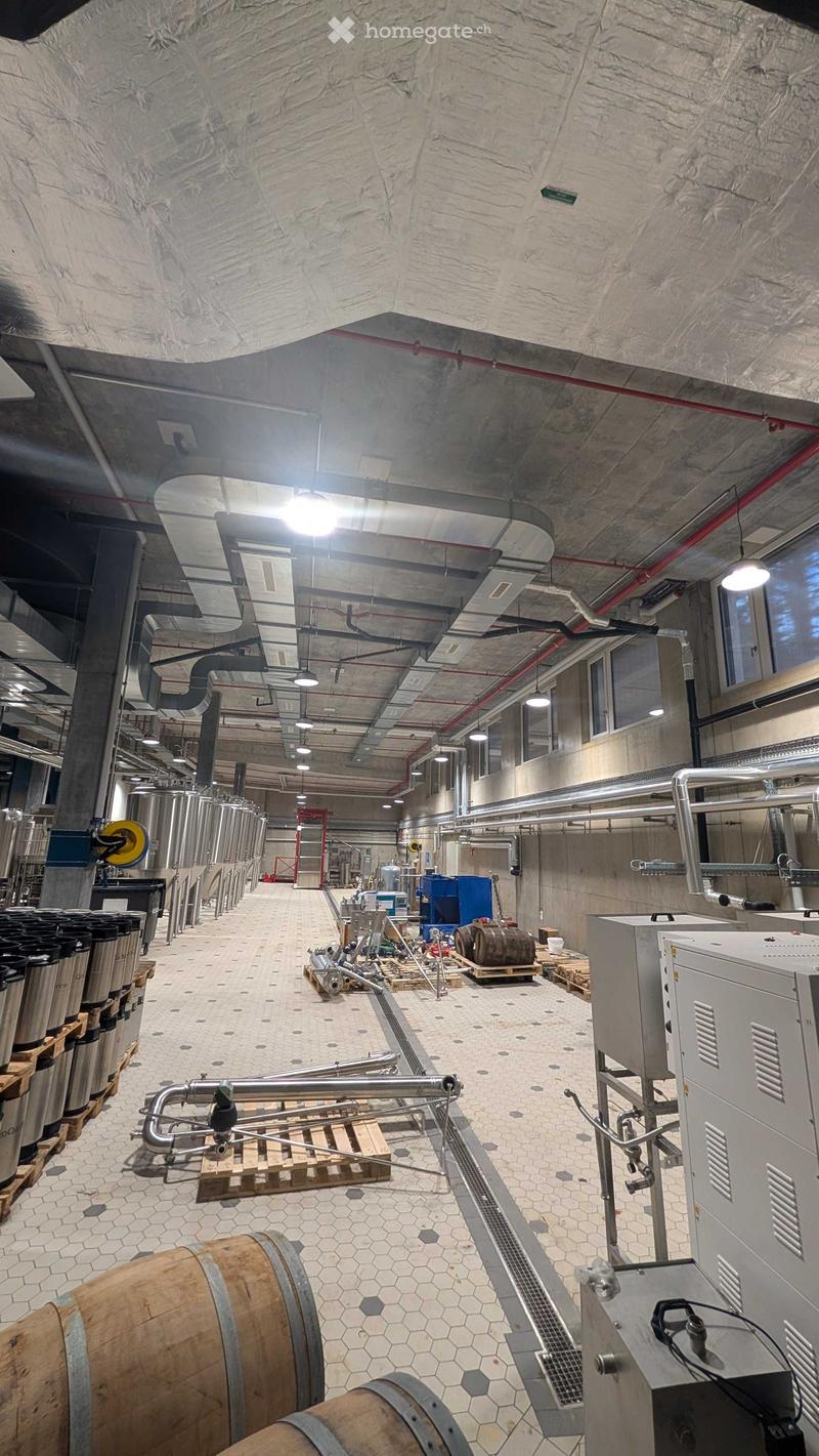 Industrial space with cylindrical tanks, pipes, barrels, and machinery, featuring a high ceiling with exposed ductwork and lighting.