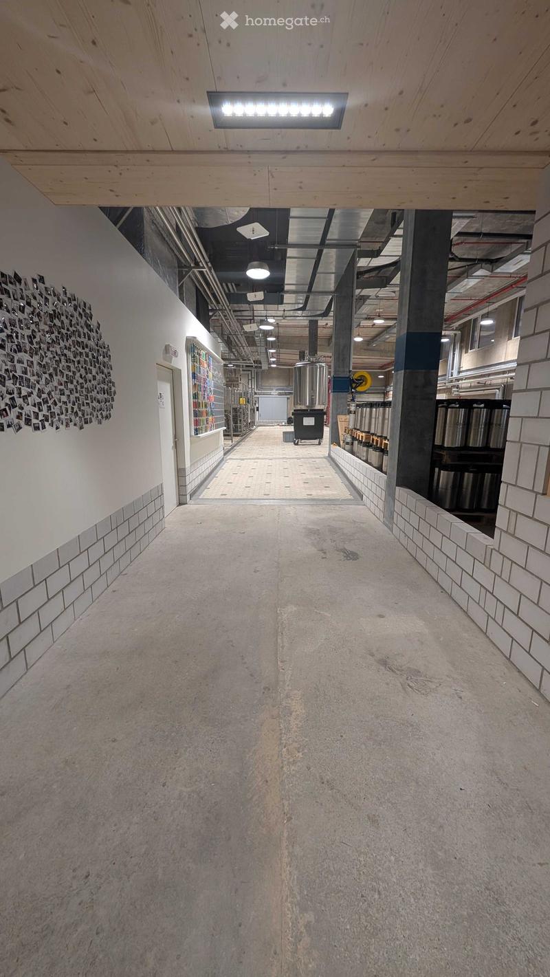 A long, industrial-style corridor with exposed pipes, concrete floors, and tiled walls. Features include a large circular photo collage on the left wall, a display shelf, and several large metal tanks in the background.