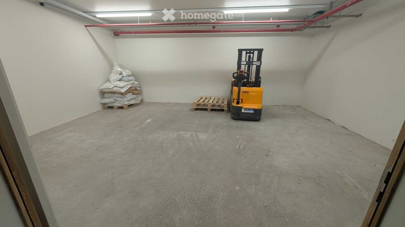 Empty room with concrete floor, white walls, and a yellow forklift in the corner. There are some pallets and bags stacked against the wall.