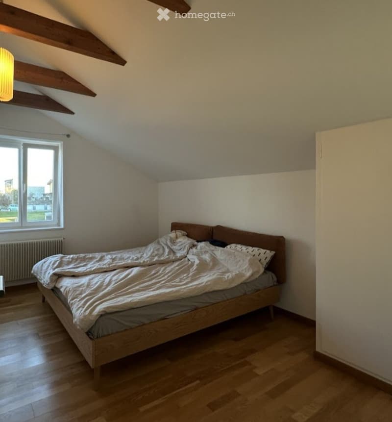 Cozy attic bedroom with wooden floors, slanted wooden ceiling beams, a large window offering a view, and a neatly made bed.