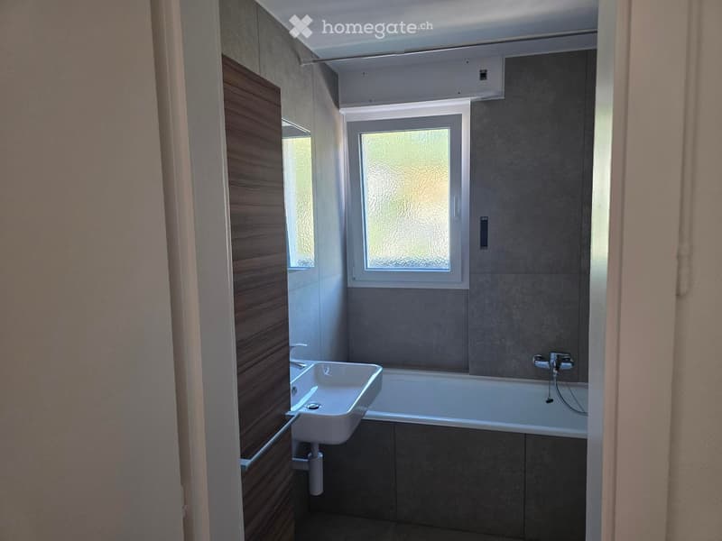 Modern bathroom with a white sink, bathtub, and frosted window. Grey tiled walls and wooden cabinetry.