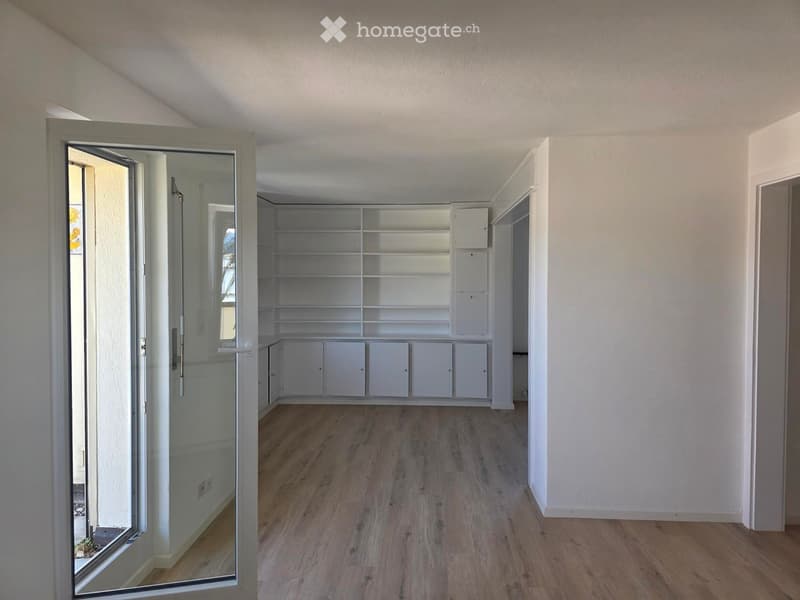 Empty room with wooden flooring, white walls, and built-in white shelving unit. The room is well-lit with natural light coming through a large window with a glass door.