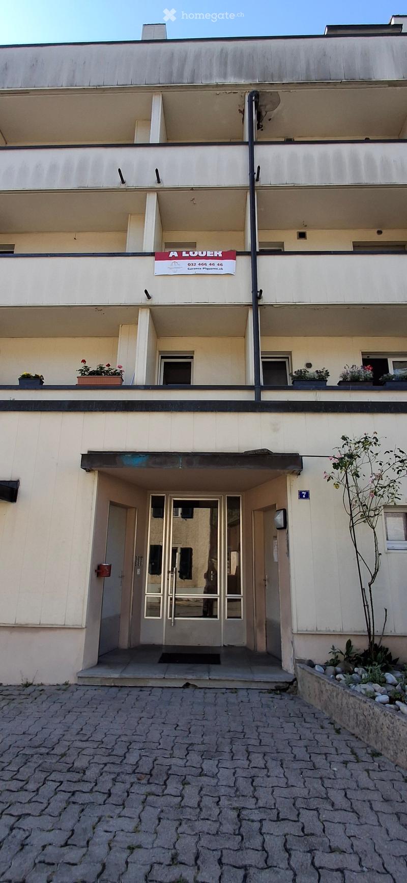 Multi-story building with a modern facade, entrance with glass doors, and potted plants on the balcony.