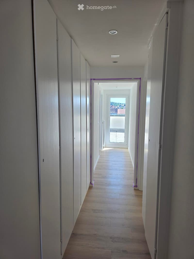 Empty hallway with wooden flooring, white walls, and recessed lighting. Doors line both sides, leading to an open area with a view of the outdoors.