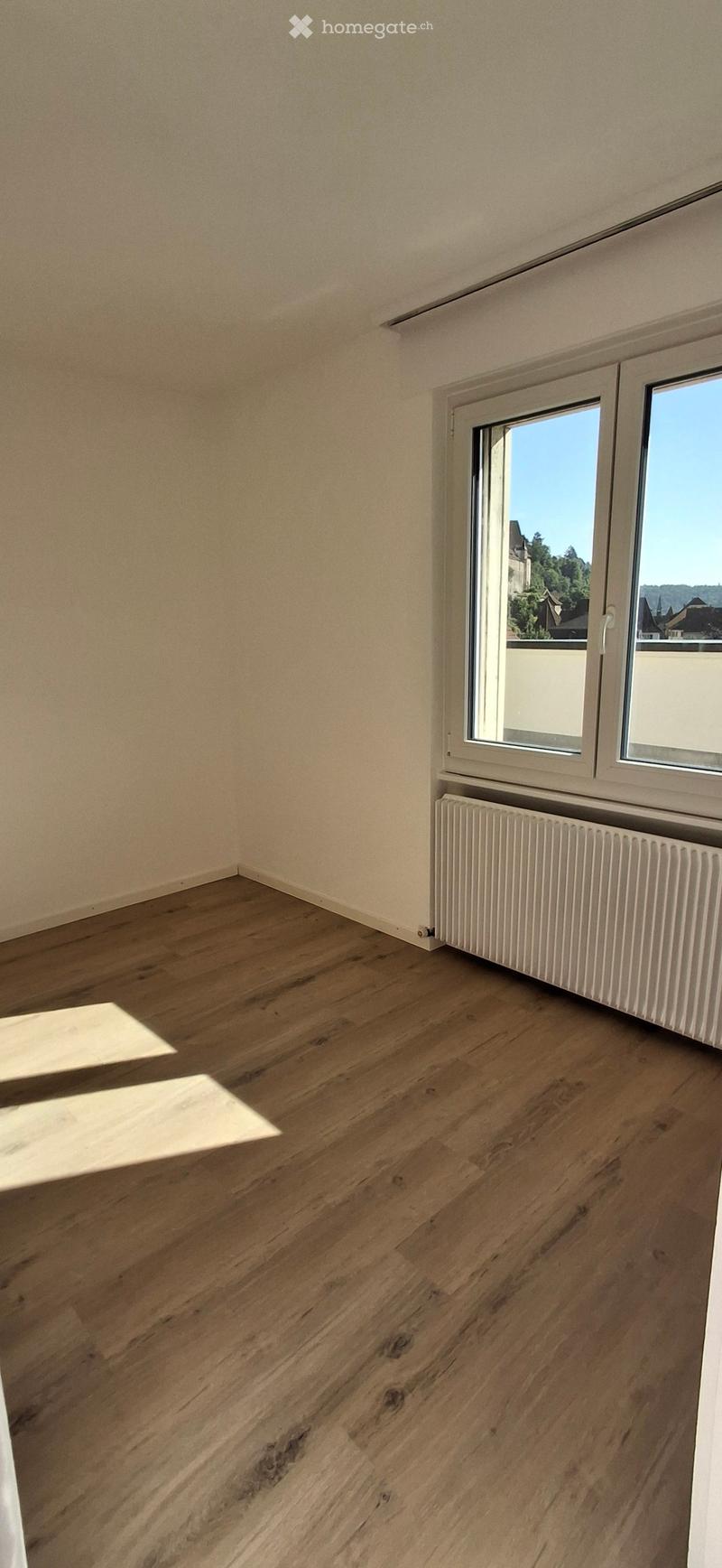 Empty room with wooden flooring, white walls, and a window with a radiator below it.
