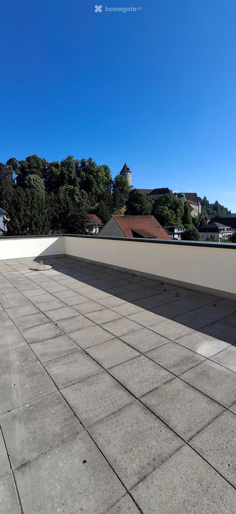 Flat rooftop terrace with concrete tiles, low white walls, and a view of surrounding buildings and greenery.