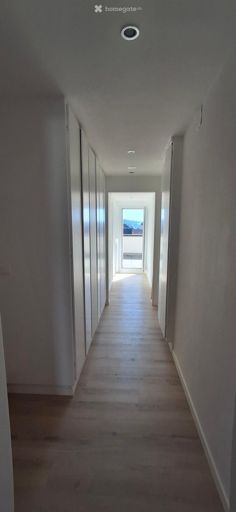 Empty hallway with light-colored walls, wooden flooring, and recessed ceiling lights. The hallway leads to an open doorway with a view of a balcony or outdoor area.