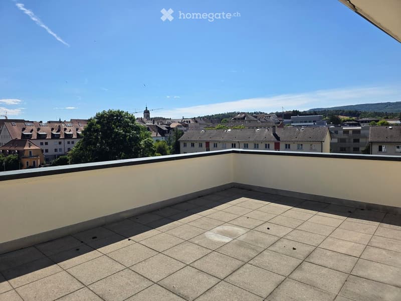 Empty rooftop terrace with tiled flooring and low parapet walls, overlooking a cityscape with buildings and trees.