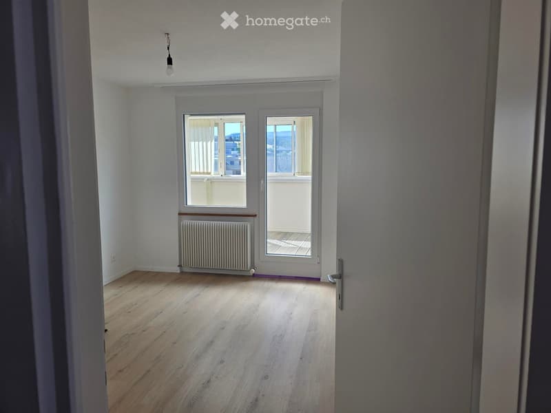 Empty room with wooden flooring, white walls, large windows, radiator, and a view of the outside area.