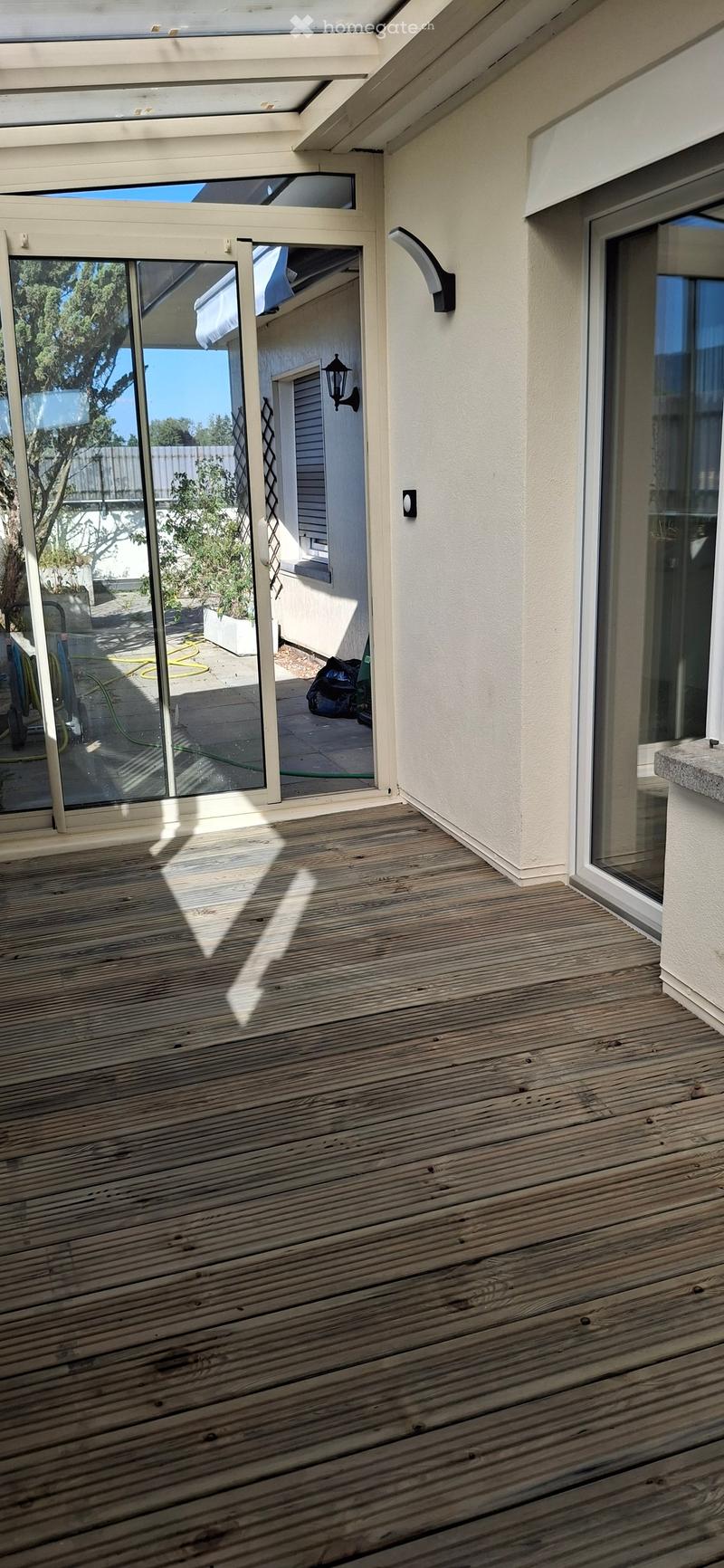 Wooden deck with large glass sliding doors, modern lighting fixture on wall, and view of outdoor area with plants and a hose.