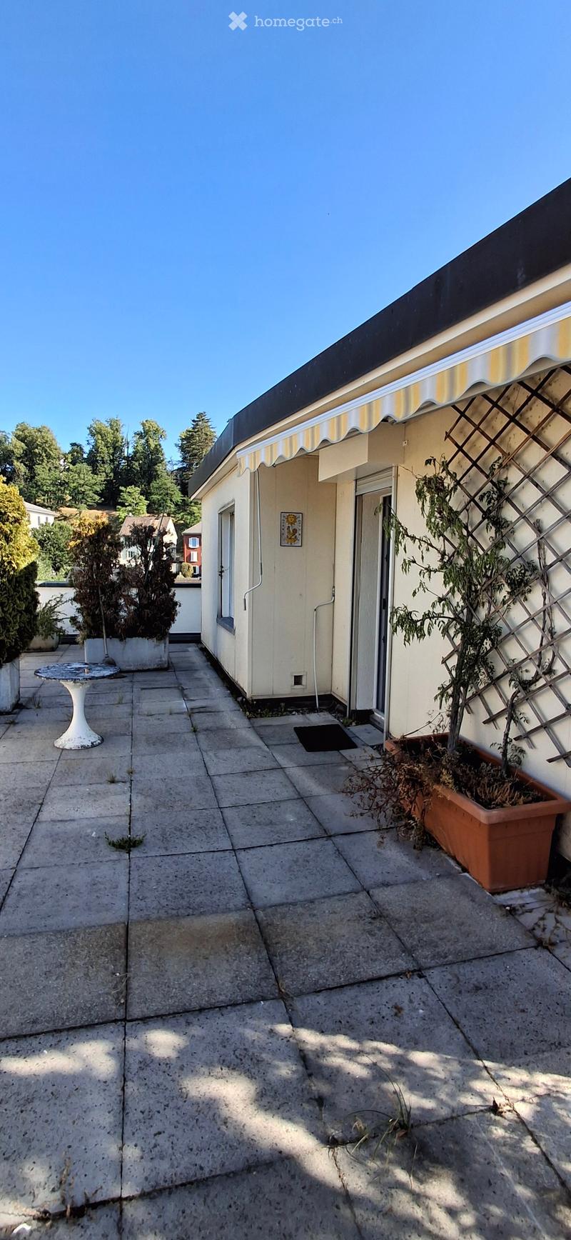 Concrete patio with a small table, potted plants, and a building entrance with a striped awning.