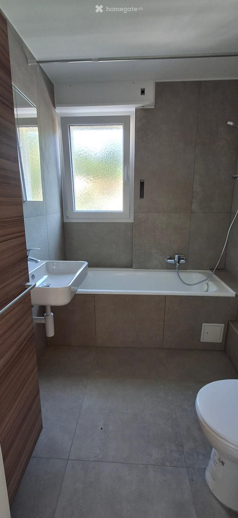 Modern bathroom with a white sink, bathtub, and toilet, featuring grey tiled walls and floor, and a frosted window above the bathtub.