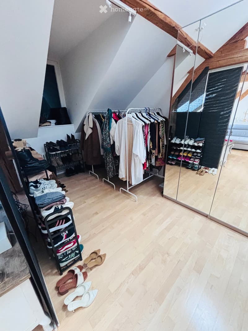 Attic room with wooden flooring, slanted wooden ceiling beams, large mirror, clothing rack, shoe racks, and neatly arranged shoes and clothing.