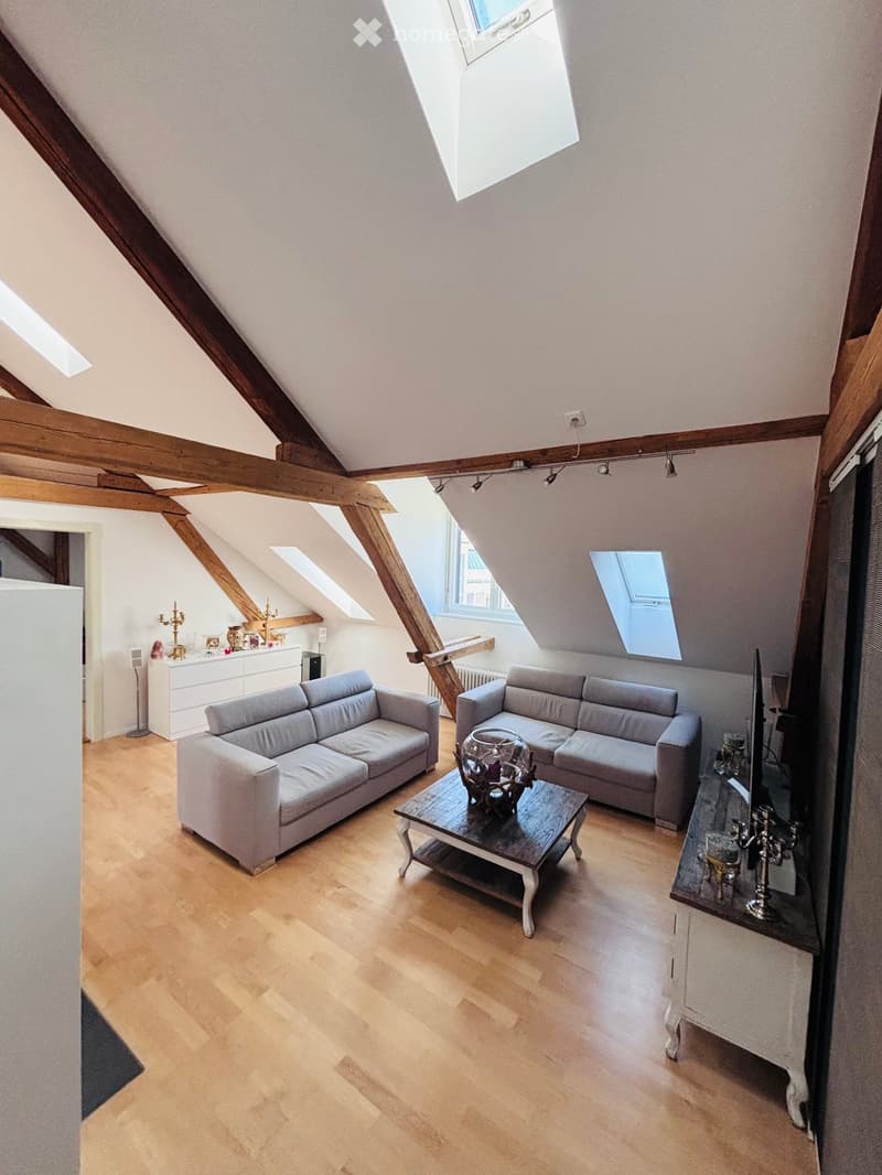 Bright living room with wooden floors, exposed wooden beams, and skylights. Features two grey sofas, a coffee table with a decorative centerpiece, and a cabinet with decorative items.