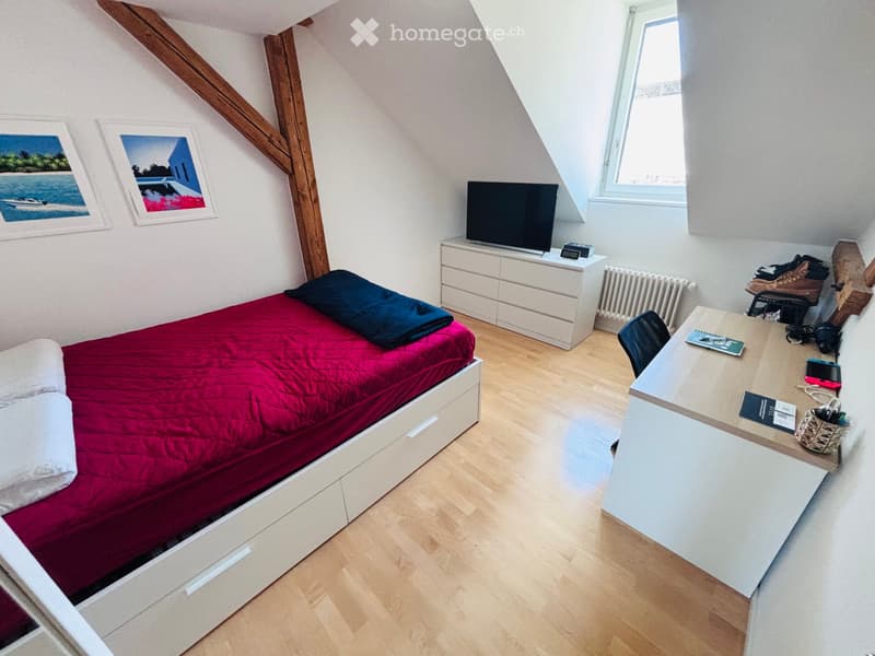 Cozy attic bedroom with wooden floors, slanted wooden beams, a bed with red bedding, a desk with a chair, a dresser with a TV, and a radiator.