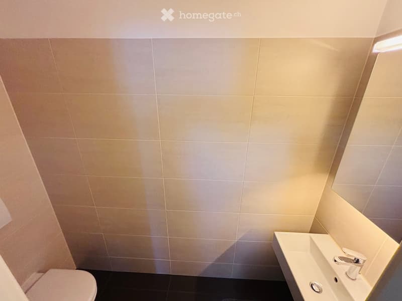 Modern bathroom with beige tiled walls, black tiled floor, white toilet, and white sink with chrome faucet.