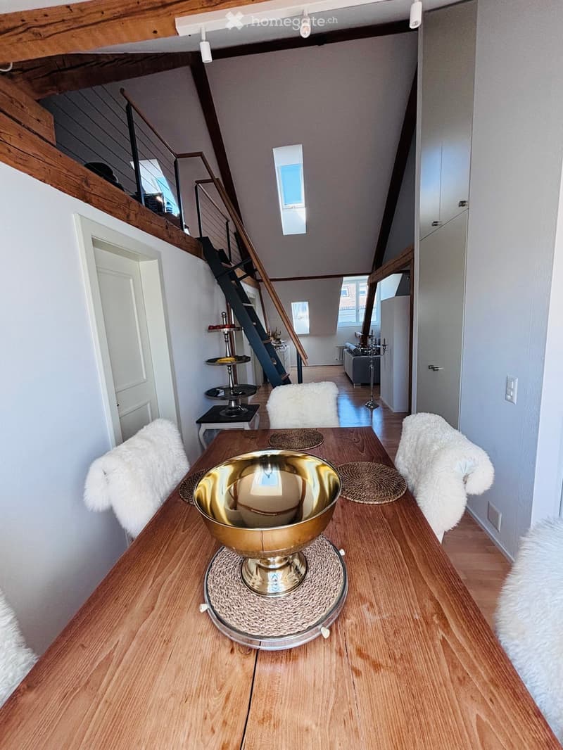 A modern hallway with a wooden staircase leading to an upper level, featuring a dining table with a large golden bowl, chairs with white fur covers, and a cat tree in the background.