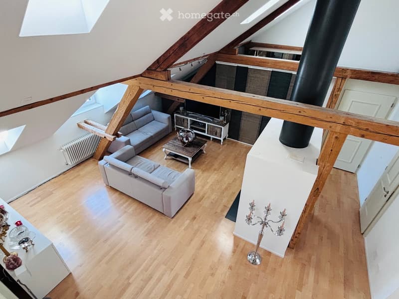 A modern living room with wooden floors, exposed wooden beams, and skylights. Features include a grey sofa, coffee table, TV stand, and decorative items.