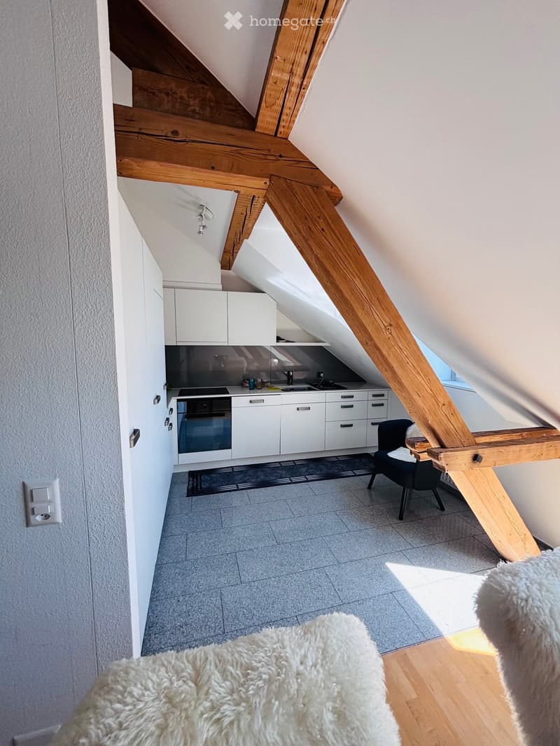 Loft-style room with exposed wooden beams, grey tiled floor, white cabinets, a small kitchen area, a black chair, and a fluffy white rug.