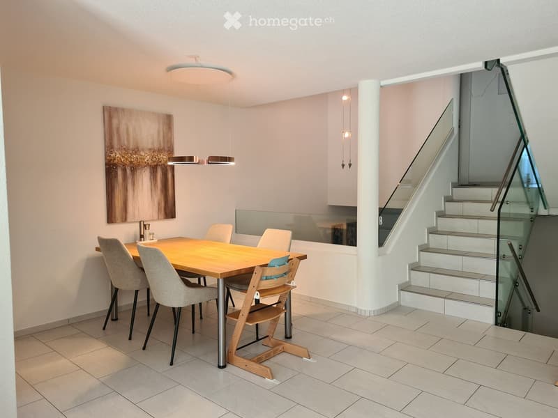 Modern dining area with a wooden table, four chairs, a baby chair, pendant lights, and an abstract art piece on the wall. A staircase with glass railings is visible in the background.
