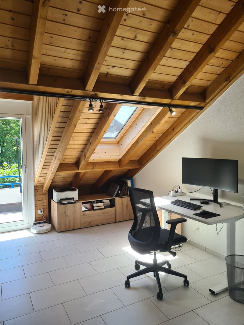 Modern attic office with wooden slanted ceilings, large skylight, white tiled floor, ergonomic chair, desk with computer setup, printer, and storage cabinet.