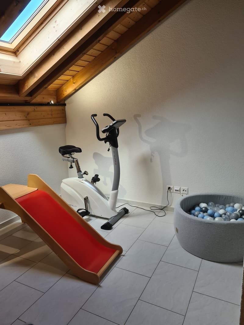 A small, well-lit room with a white exercise bike, a wooden slide, and a round ball pit filled with colorful balls.