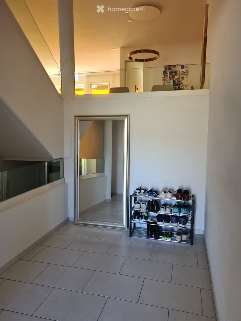 Modern hallway with tiled floor, large mirror, shoe rack, and a view to another room with a staircase and a round light fixture.