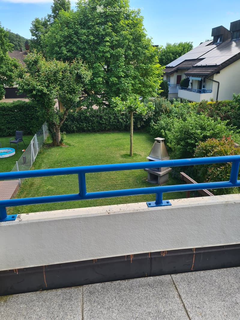 Balcony overlooking a well-maintained garden with a swimming pool, lawn, trees, and a neighboring house with a balcony.