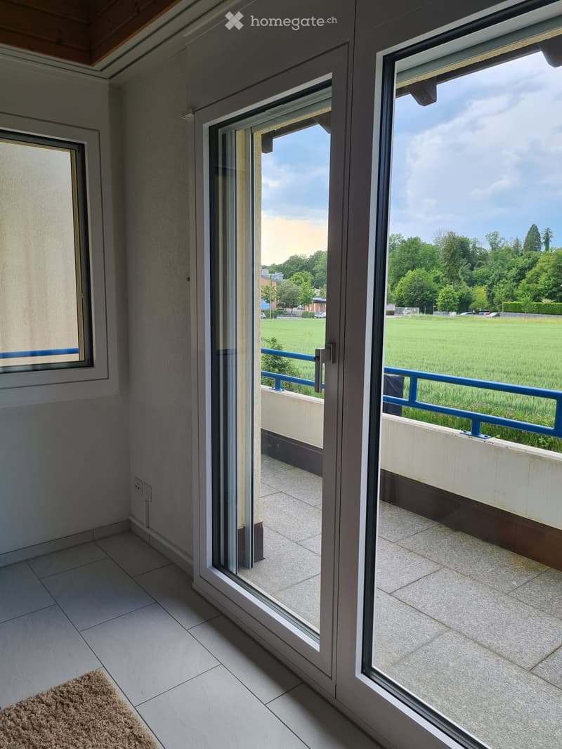 Balcony with large sliding glass doors, tiled floor, and a view of a green field and trees.