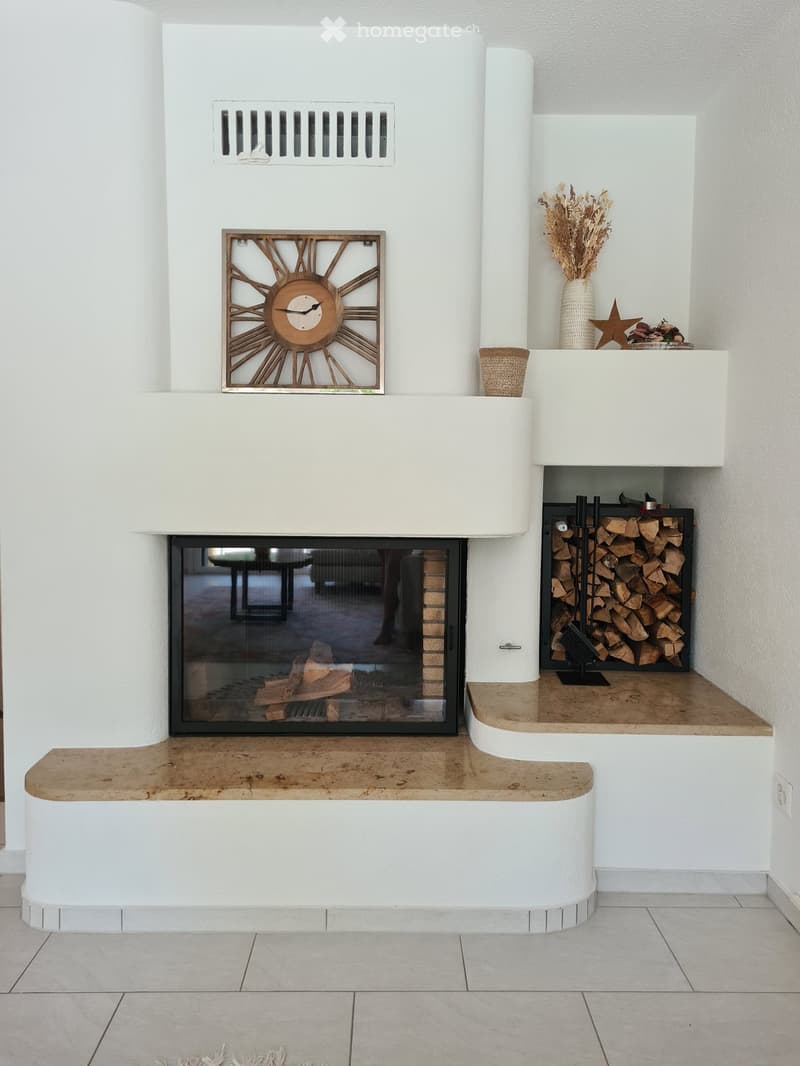 Modern fireplace with a decorative clock above, a vase with dried flowers, and a stack of firewood to the side.