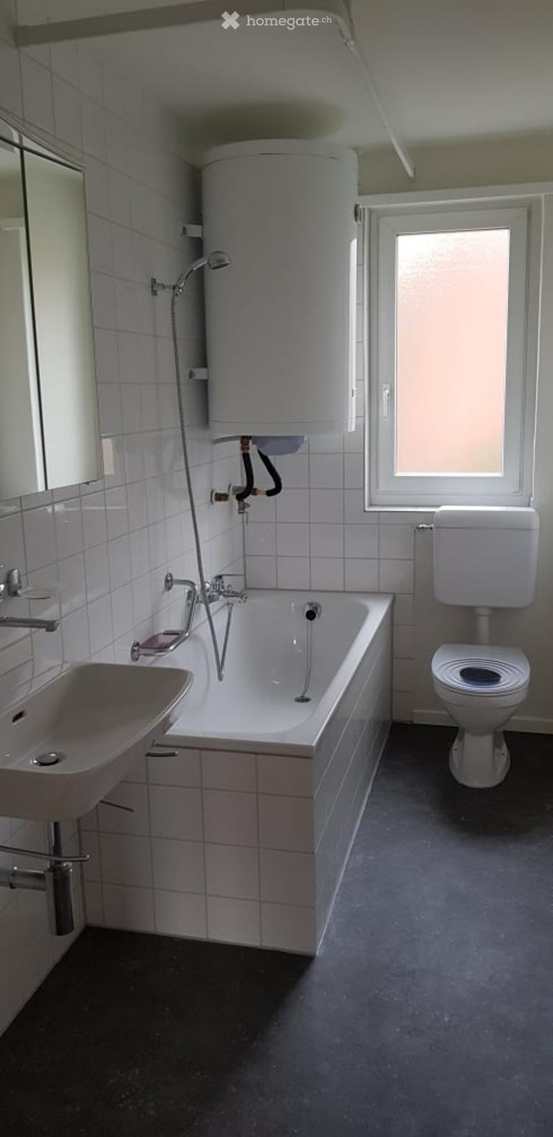 Modern bathroom with white tiles, a bathtub, toilet, sink, mirror, and a window providing natural light.