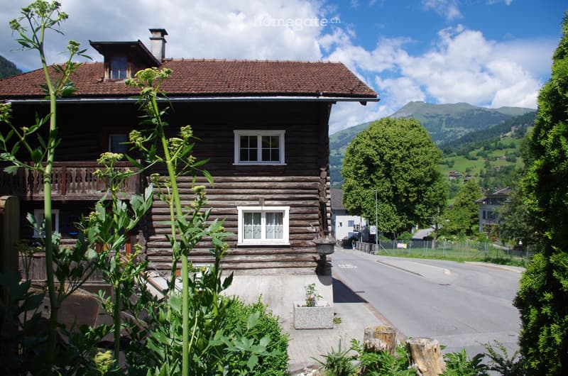 Traditional wooden house with a sloped roof, multiple windows, and a small balcony, surrounded by greenery and a road.