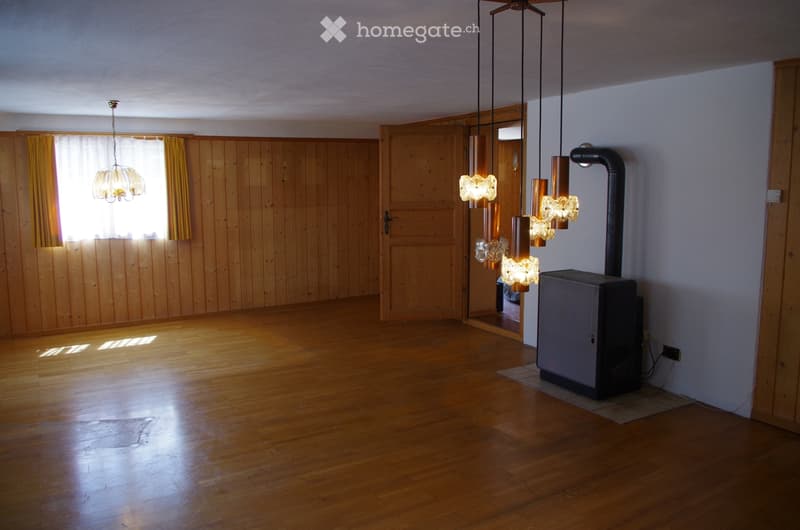 Empty room with wooden walls and floor, large window with yellow curtains, modern hanging light fixtures, and a black wood-burning stove with a chimney pipe.