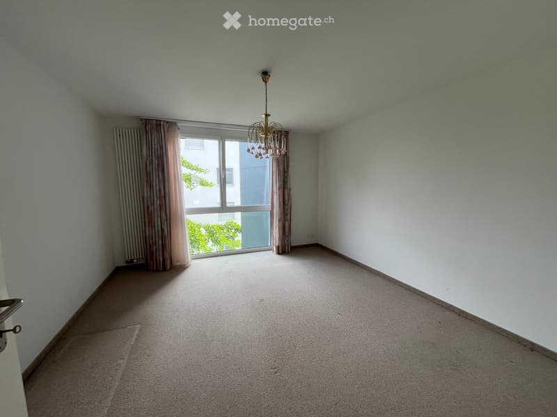Empty room with light-colored walls, carpeted floor, large window with curtains, and a chandelier hanging from the ceiling.