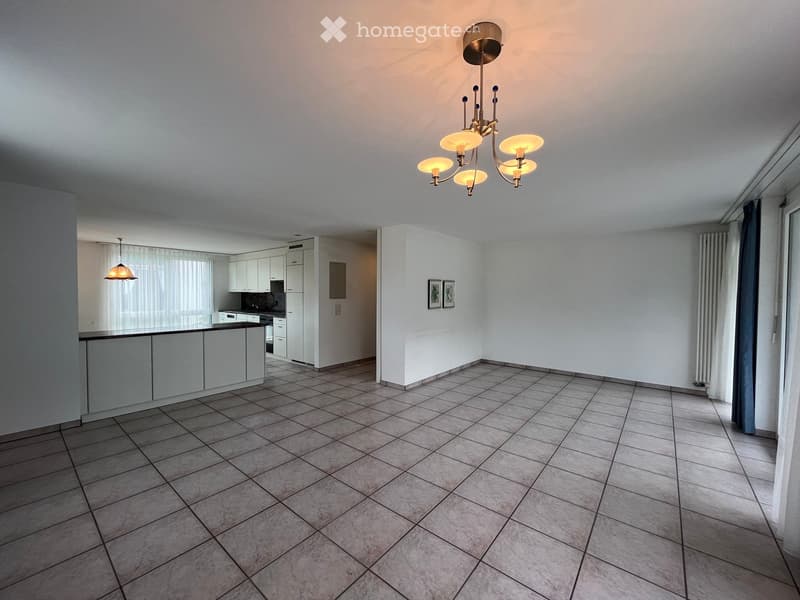 Open-plan living and dining area with tiled flooring, white kitchen cabinets, a central island, and a chandelier. The space is bright with large windows covered by curtains and includes a radiator and picture frames on the wall.
