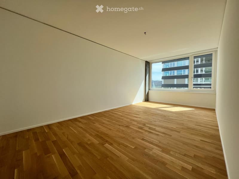 Empty room with wooden flooring and white walls, featuring large windows with blinds and a view of neighboring buildings.