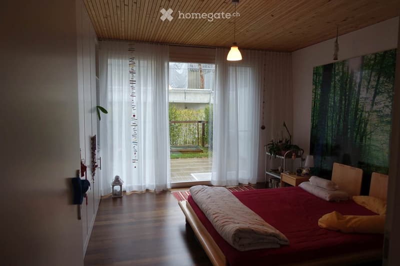 red bed, white sheets, red and yellow pillows, wooden bed frame, wooden floor, glass doors to balcony, plants in window, shelf with plant and lamp