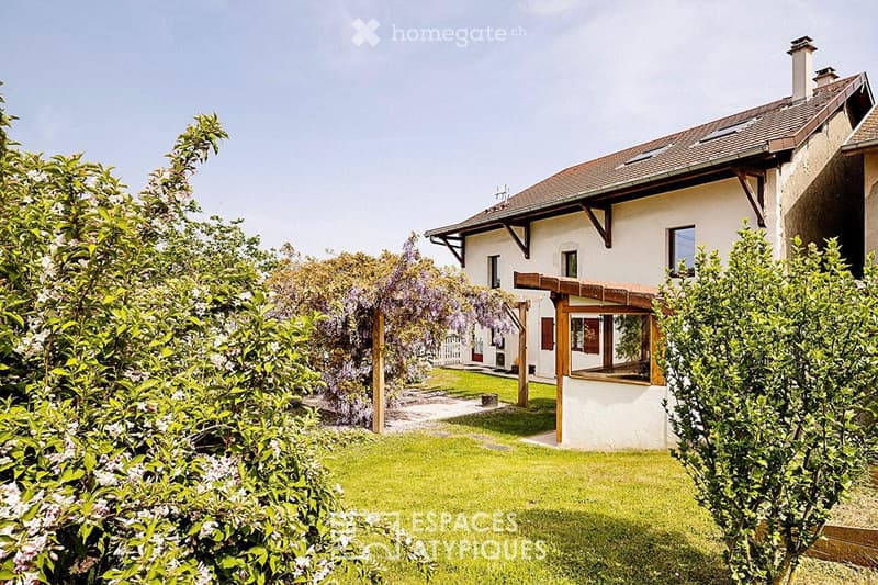 A two-story house with a tiled roof, white exterior, and a covered wooden porch. The house is surrounded by a well-maintained garden with lush greenery and flowering shrubs.