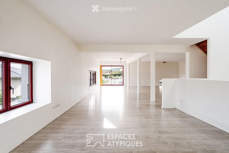 Bright, open corridor with white walls, wooden flooring, and large windows allowing natural light. Features a hanging light fixture and a view of an outdoor area through a glass door.