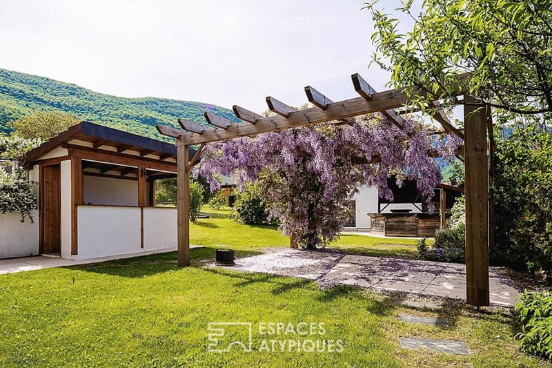 A well-maintained garden with a wooden pergola draped with purple wisteria, a small wooden structure, a stone pathway, and a grassy lawn.