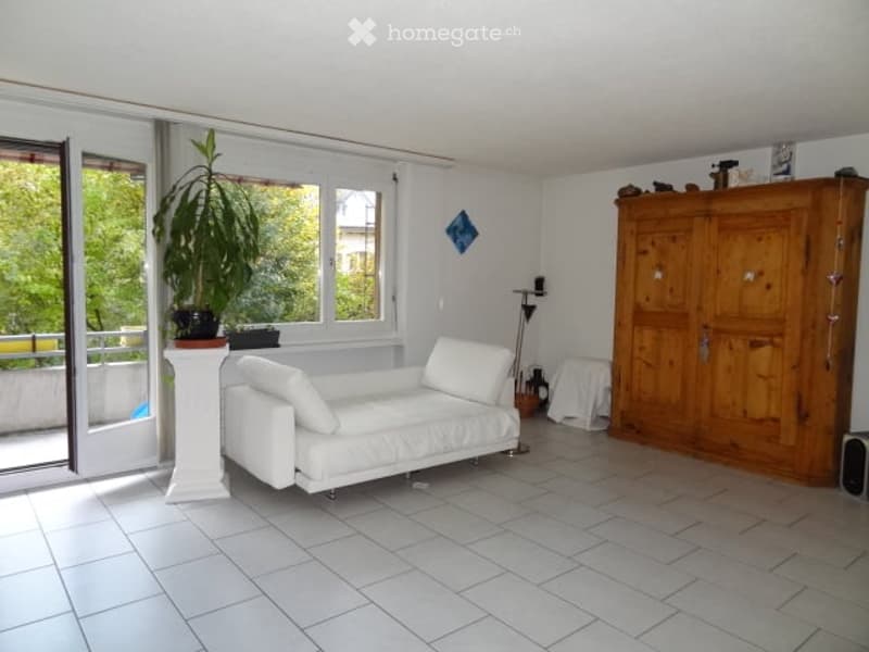 Bright living room with white tiled flooring, large windows, a white sofa, a wooden cabinet, and a potted plant near the balcony entrance.